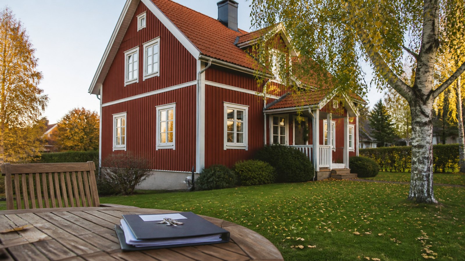 Röd trävilla med vita knutar och en pärm med husnycklar på trädgårdsbordet en höstdag i Sverige